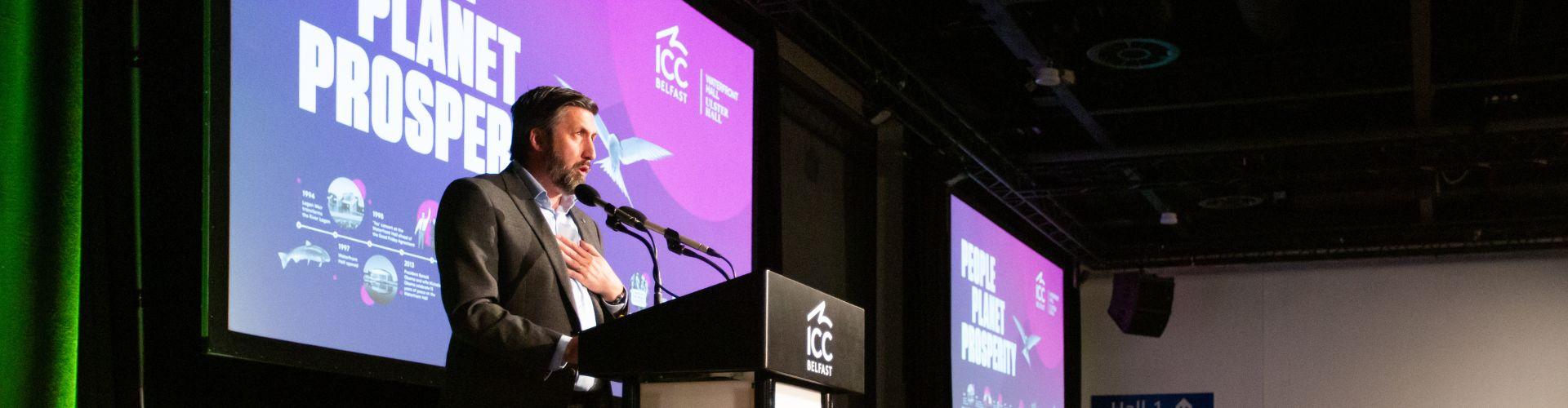 Man stands at lectern during Sustain Exchange conference for keynote speech