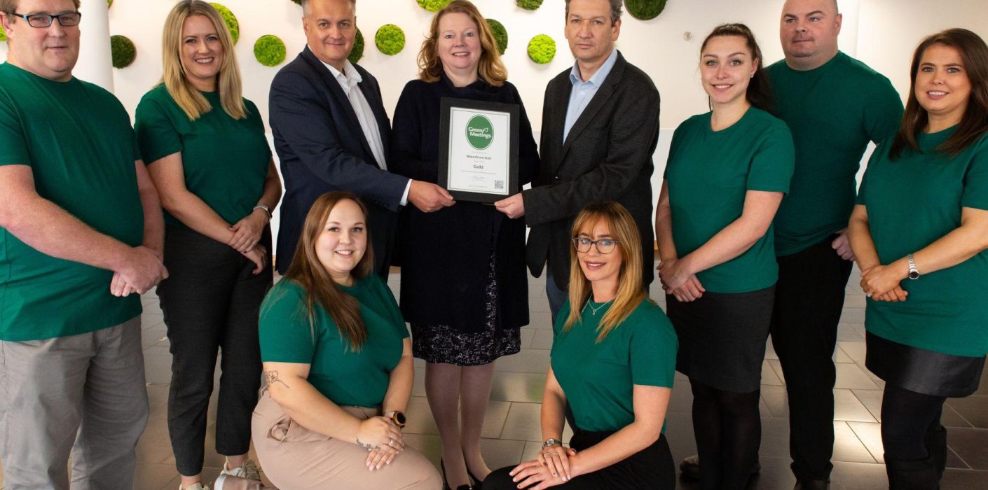 Ten adults standing facing the camera in foyer with centre three people holding picture frame with award certificate
