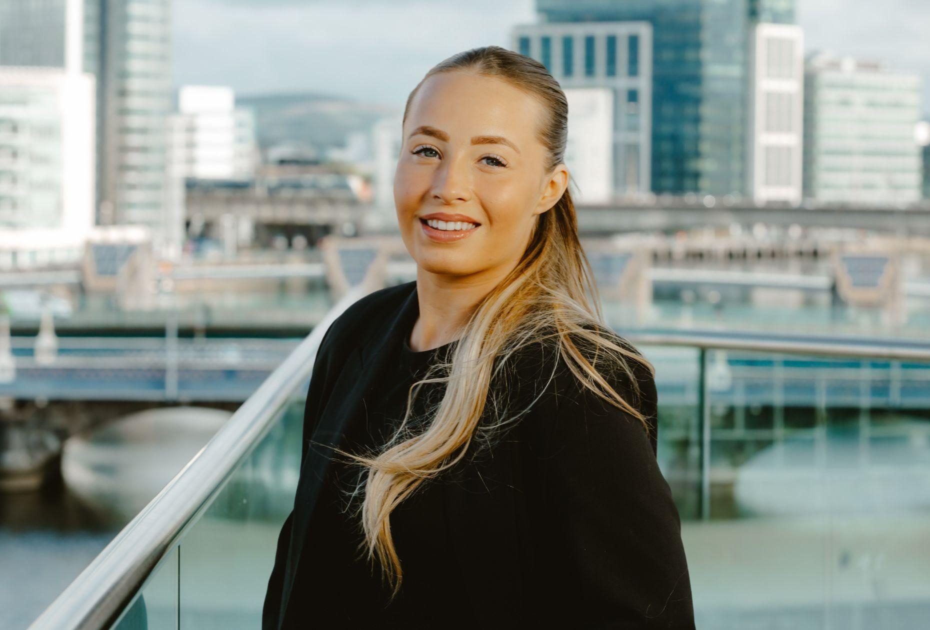 Woman standing smiling for a photo with a backdrop of Belfast city skyline in the distance