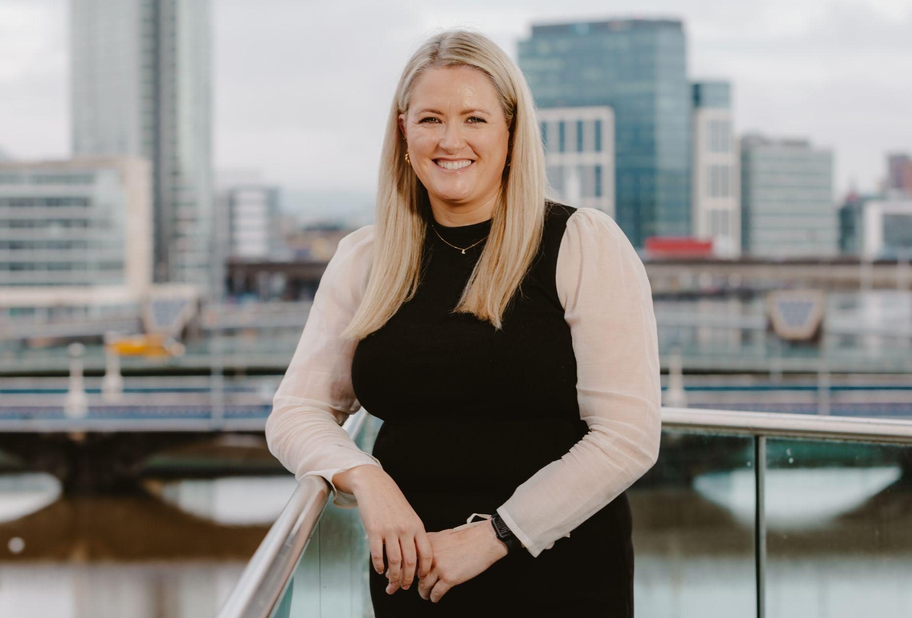 Woman standing smiling for a photo with a backdrop of Belfast city skyline in the distance