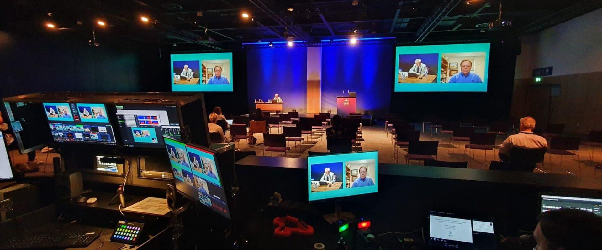 Hall 2 filled with delegates facing a stage. There are multiple monitors for video call purposes.