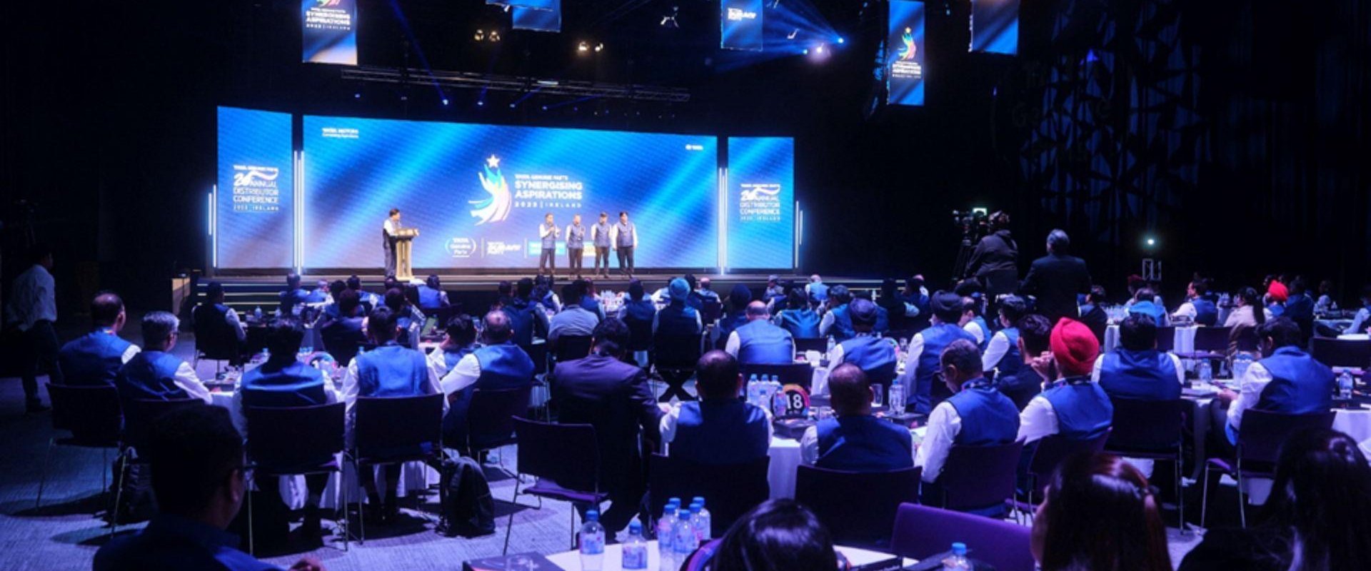 Delegates seated at round tables for the Tata Genuine Parts for their Annual Distributor Conference. They are facing a stage with four members stood to the right and one at a podium on the left. The large screen behind them reads, 'Tata Genuine Parts, Synergising Aspirations, 2023, Ireland.'