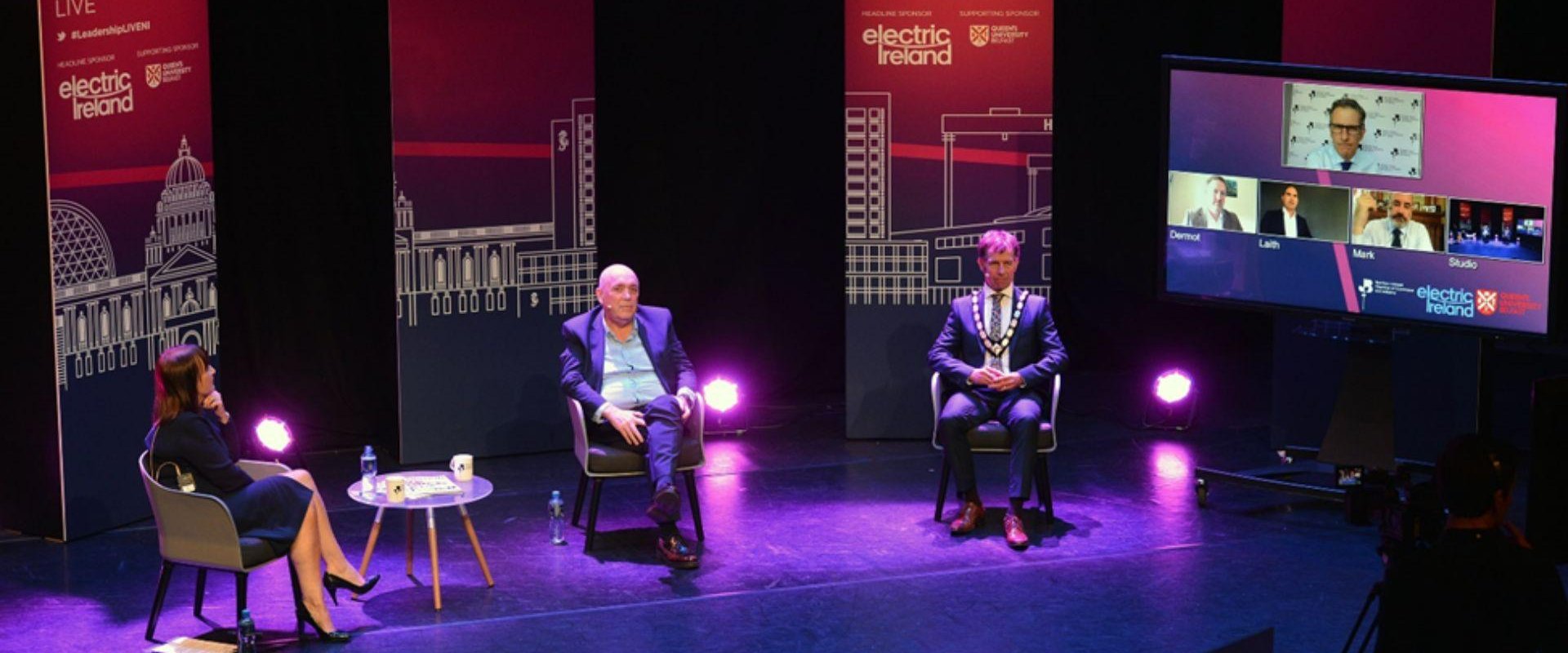 Three people seated on a stage for a discussion. Banners are displayed at either side of the people involved and two the right of them is a large screen with other members on a video call to join the hybrid event.
