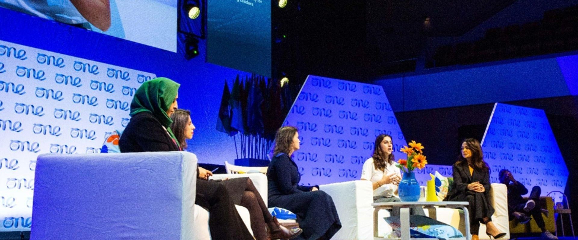 Five female delegates seated on the main auditorium stage during the one young word event with the company branding distributed behind them, alongside a large screen that shows close up shots of each delegate.