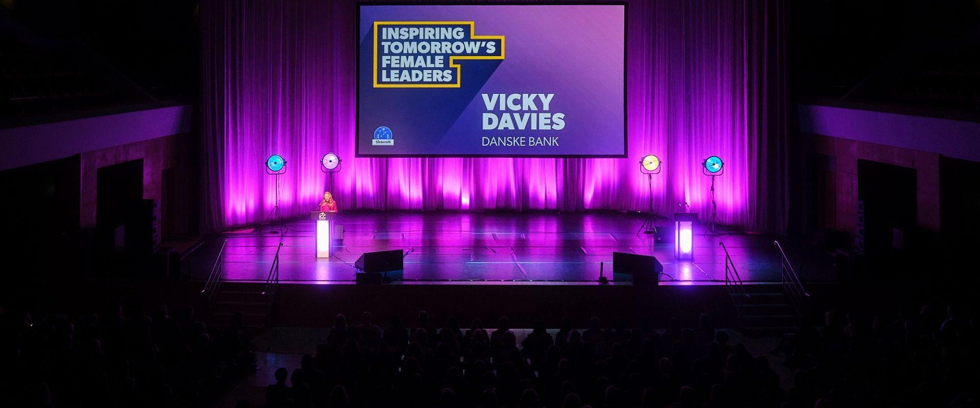 Main Auditorium set up for the SistersIN celebration event. Vicky Davies is stood at a podium delivering a presentation to the attendees and is stood in front of a large digital screen that displays her name and reads, ' Inspiring tomorrow's female leaders'