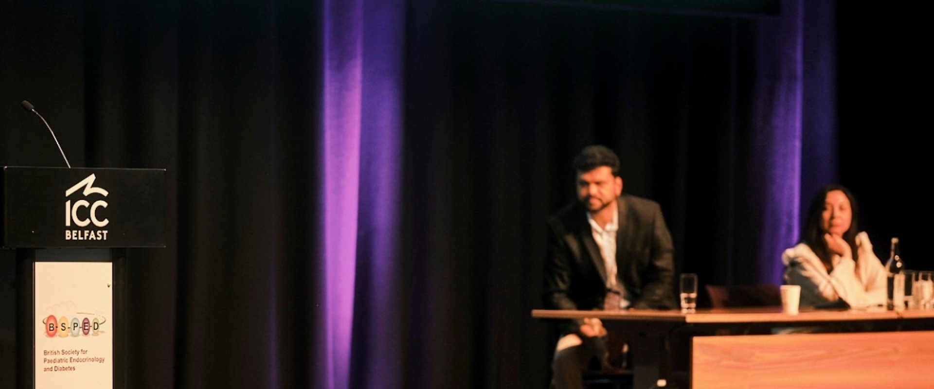 Tabitha Randell, president of BSPED, addresses people from podium on stage, with two members of her team in the background sitting at a table.