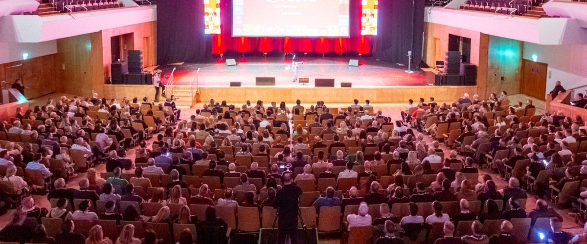 Hundreds of employees sit in Main Auditorium at ICC Belfast for the annual general meeting AGM. Main stage has large digital screen which is illuminated red with the words 'Evolve' on show.