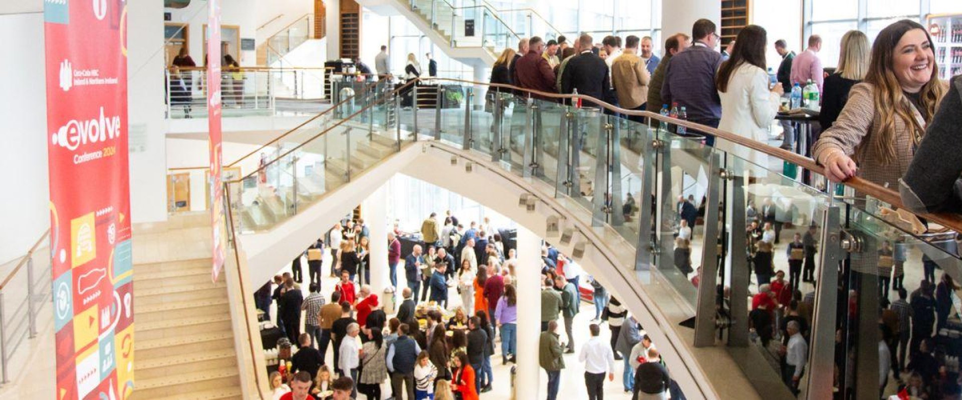 Delegates gather across multiple levels in a building with a focus on a group of women with their arms on the railings and laughing. There is a hanging red banner hanging over a staircase on the left hand side.