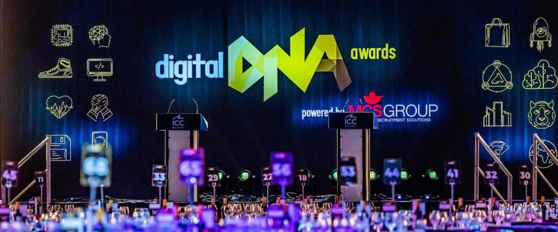 Hall 1 in ICC Belfast setup for Digital DNA 2022. There is a screen lit up with the Digital DNA yellow and white logo as well as podiums on stage and tables set up for delegates.