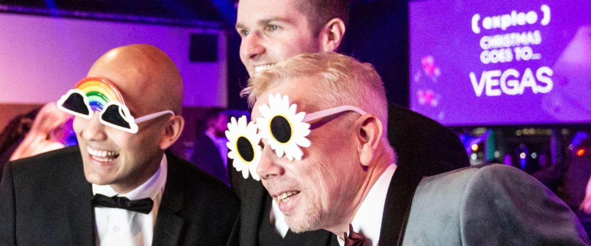 Three smiling men are getting a picture taken wearing prop sunglasses. Each man is wearing a tuxedo and in the background a purple screen is visible with the words 'Expleo Christmas Vegas Party'