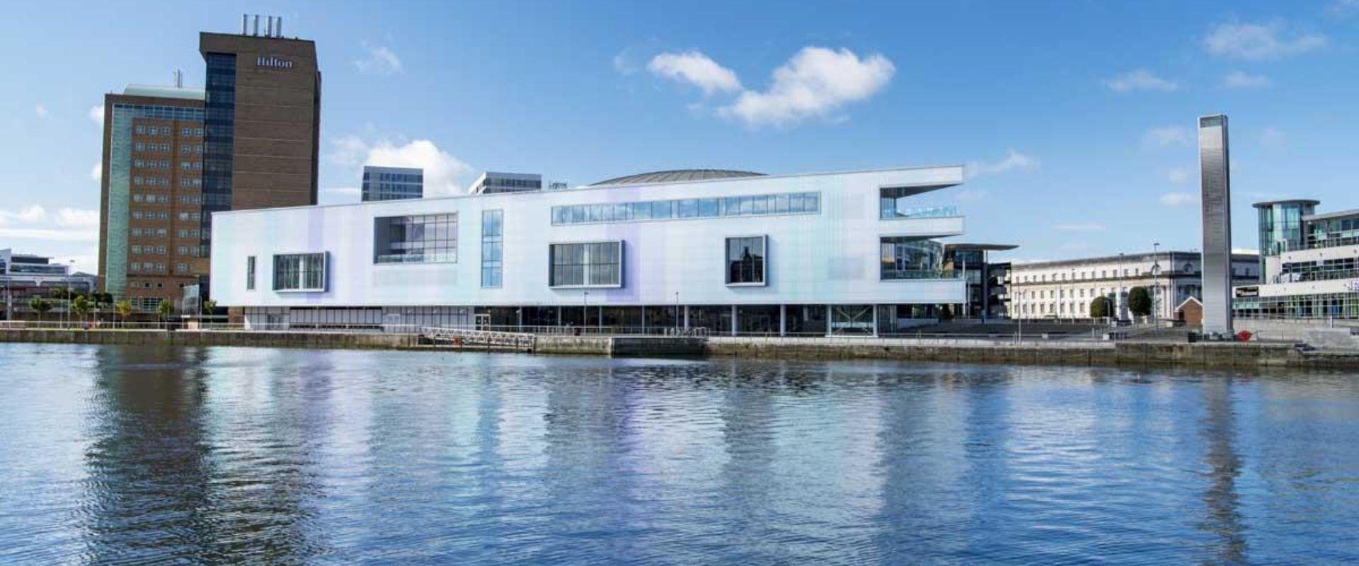 Image taken from across the water showing exterior image of white building along the water's edge with Hilton hotel showing in the background.