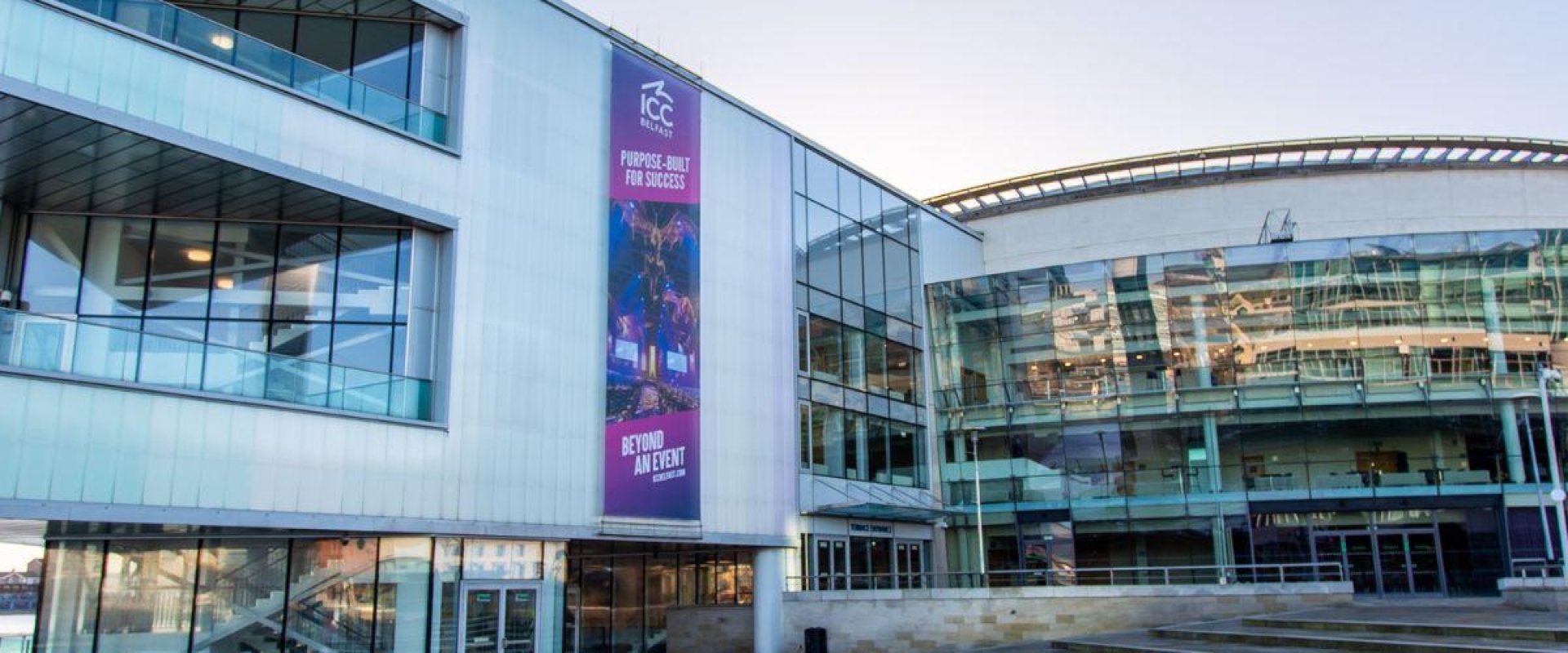 Image is a view of the exterior of ICC Belfast showing the Riverside entrance and the bars.
