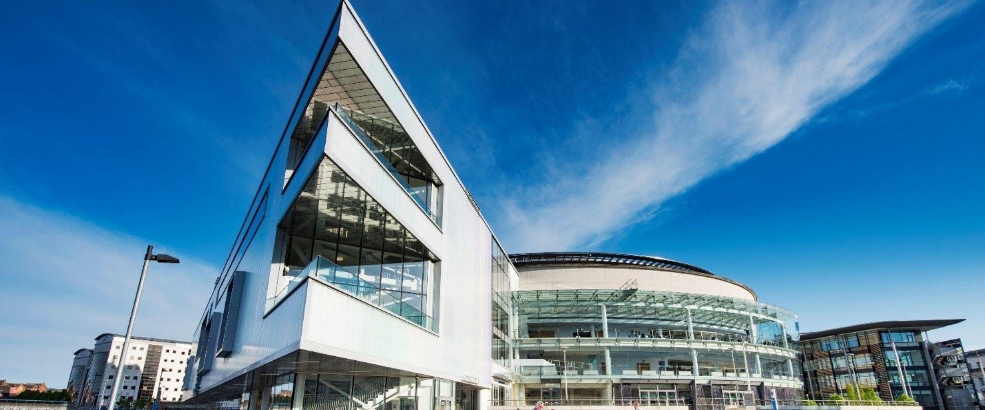 Image of the exterior of ICC Belfast taken in the sunshine in front of the entrance beside the river with individuals on the steps outside the building.