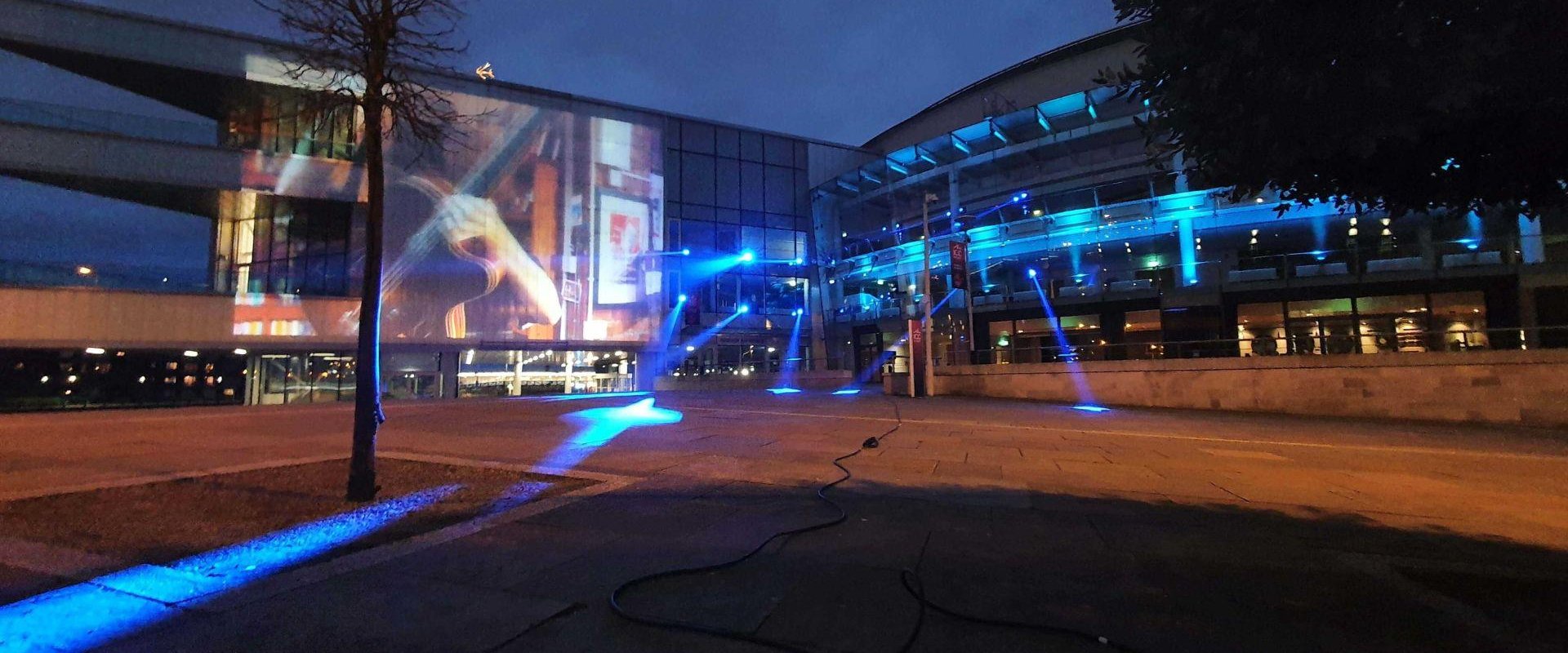 The exterior of ICC Belfast at night with a large image projected on the wall of the Riverside extension. Blue spotlights are also shining onto the building.