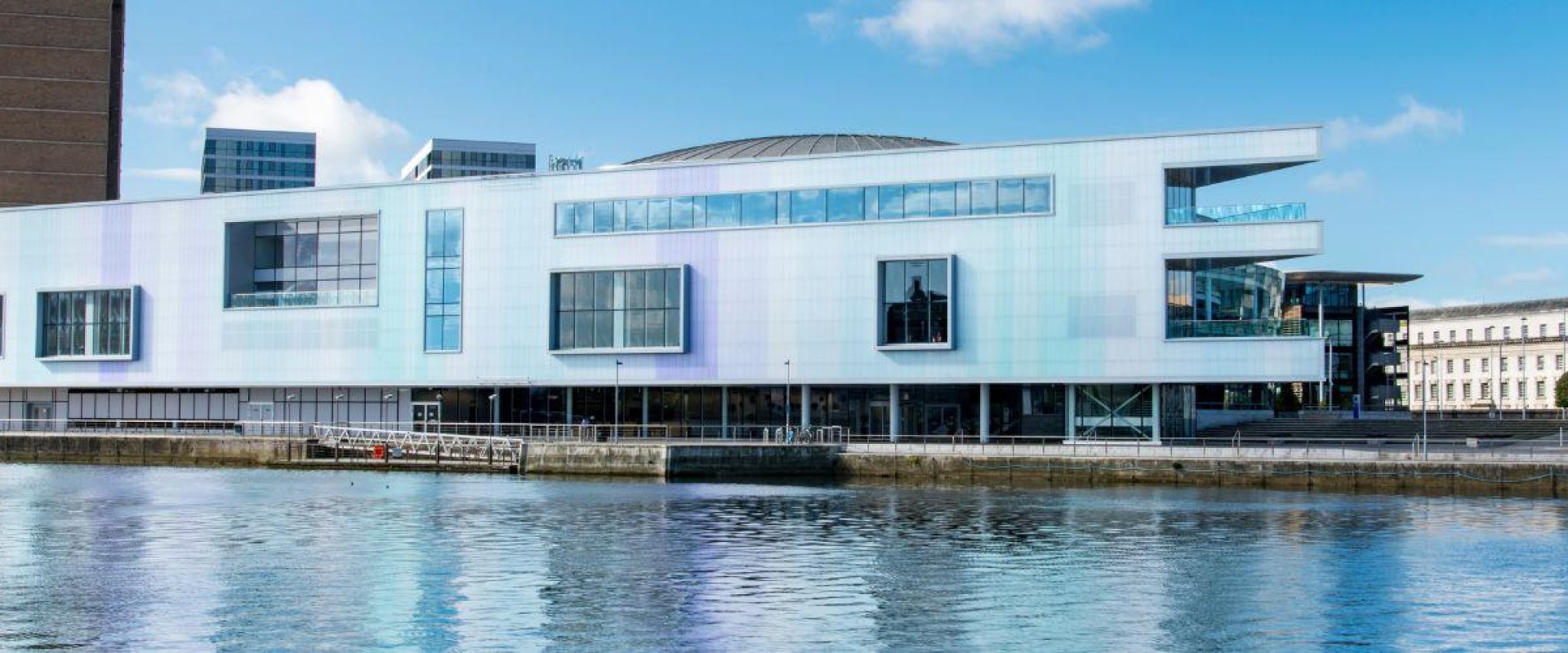 White horizontal building situated along a river with multiple windows. ICC Belfast venue hire for conferences in the UK and Ireland.