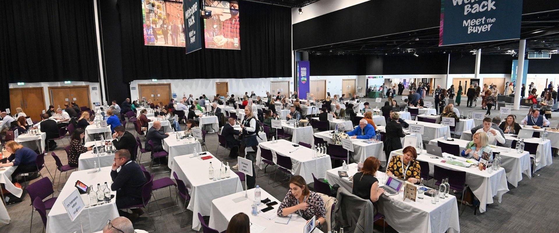 Delegates seated in Hall 1 at rectangle tables of four with a large banner at the right side of the image that reads 'welcome back, meet the buyer'