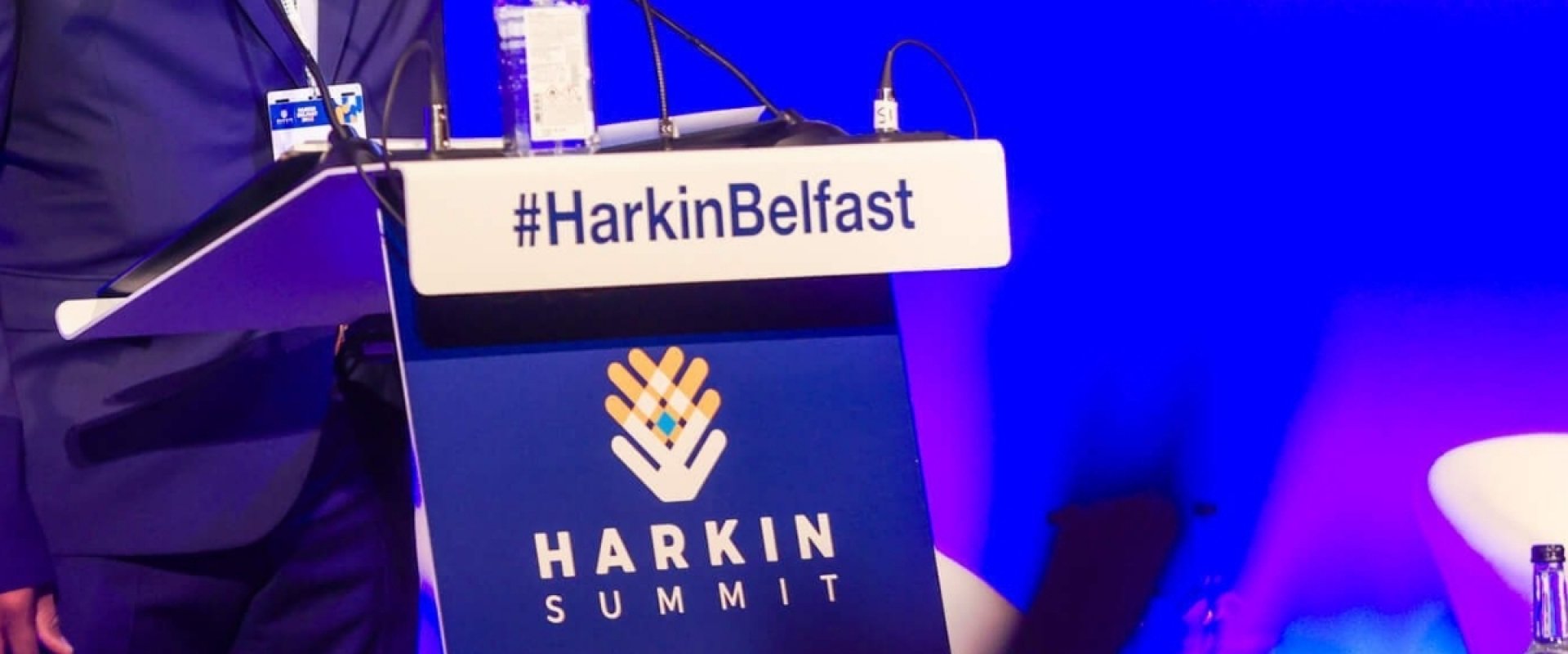Dr Joseph Jones, Executive Director at Harkin Institute waving on stage to the audience as he stands at a podiym with the Harkin Summit branding on it.