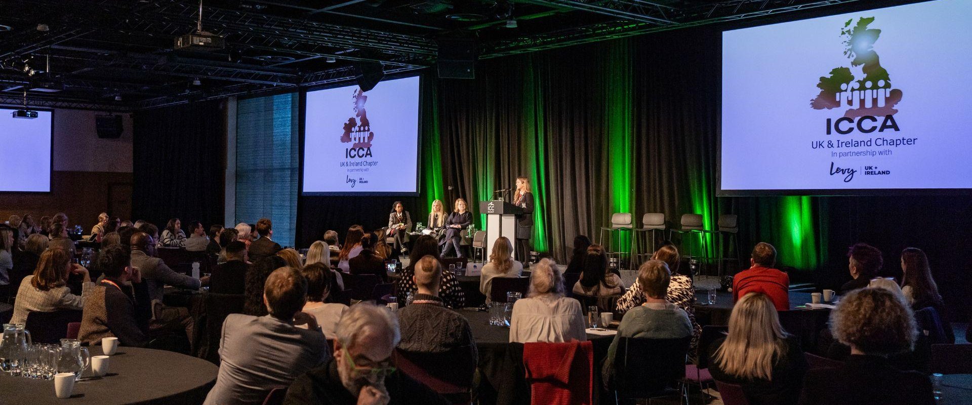The audience is paying attention to a SistersIN pupil who is giving a speech on stage, there are also digital screens in the background with the ICCA logo and green lights displayed.