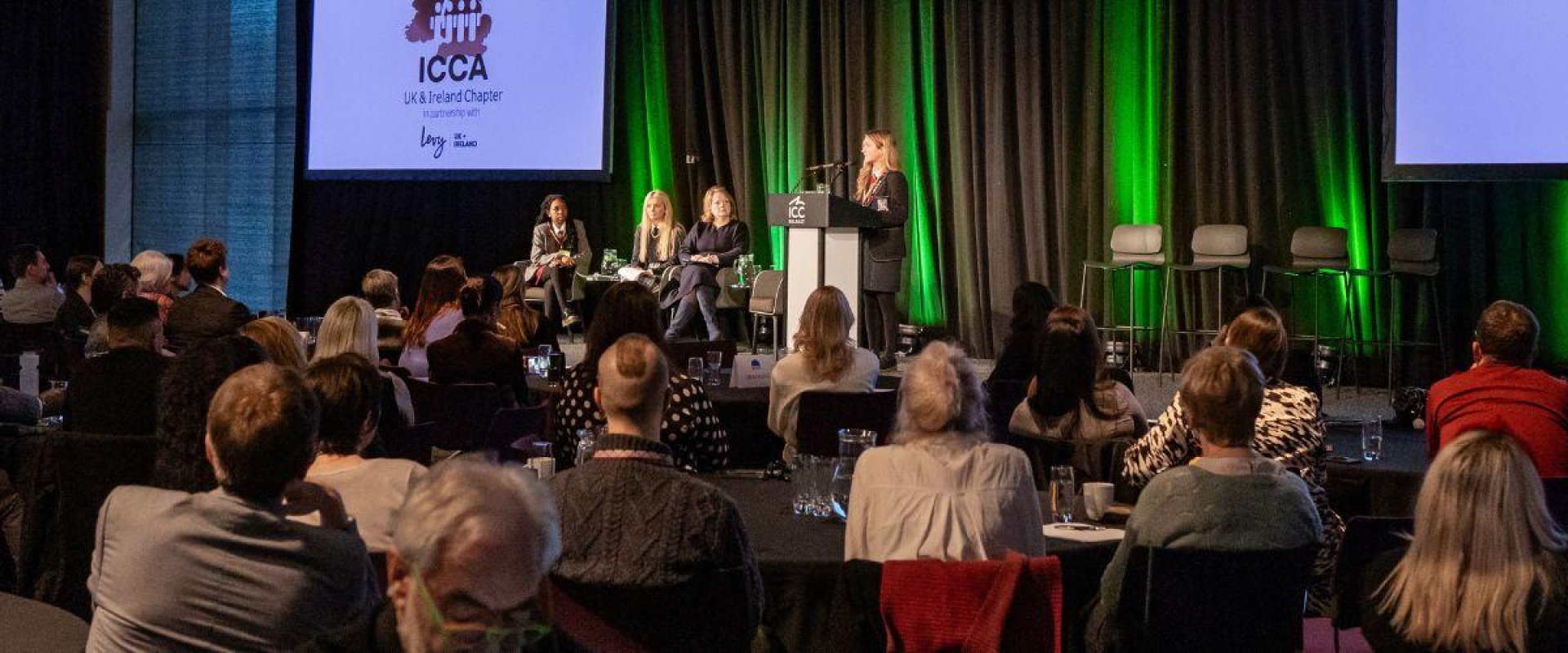 Students present on stage at the ICCA UK & Ireland Chapter to a packed hall full of people sitting at round tables on chairs