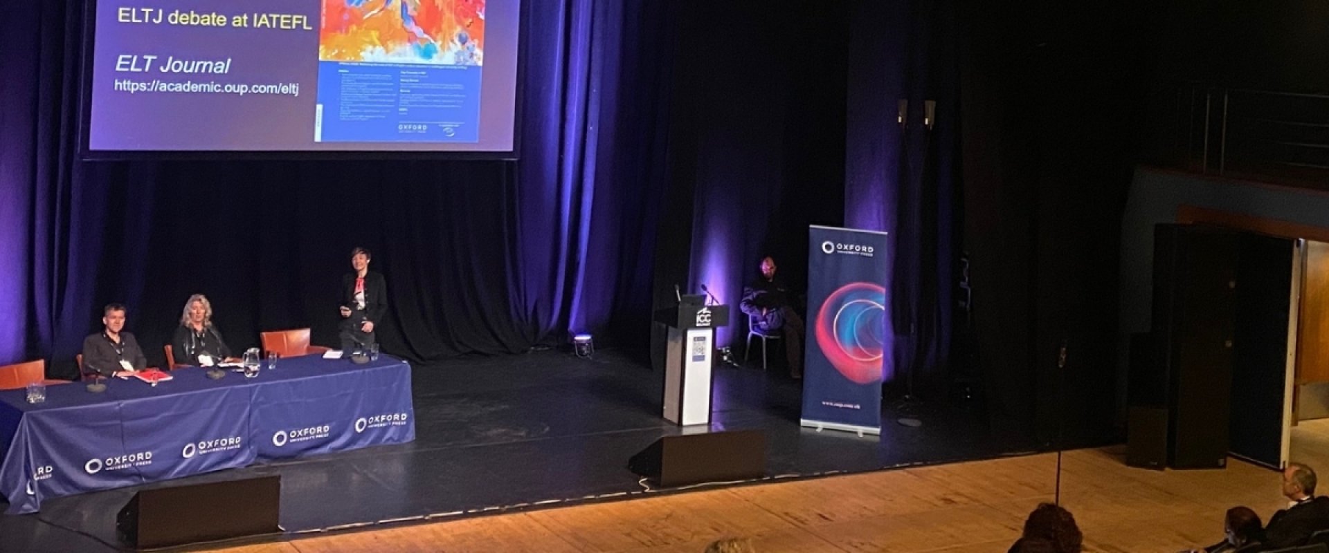 Photo shows a conference being held in the studio with a panel of individuals on stage with a presentation on screen in the background. Delegates are seated in front of the stage.