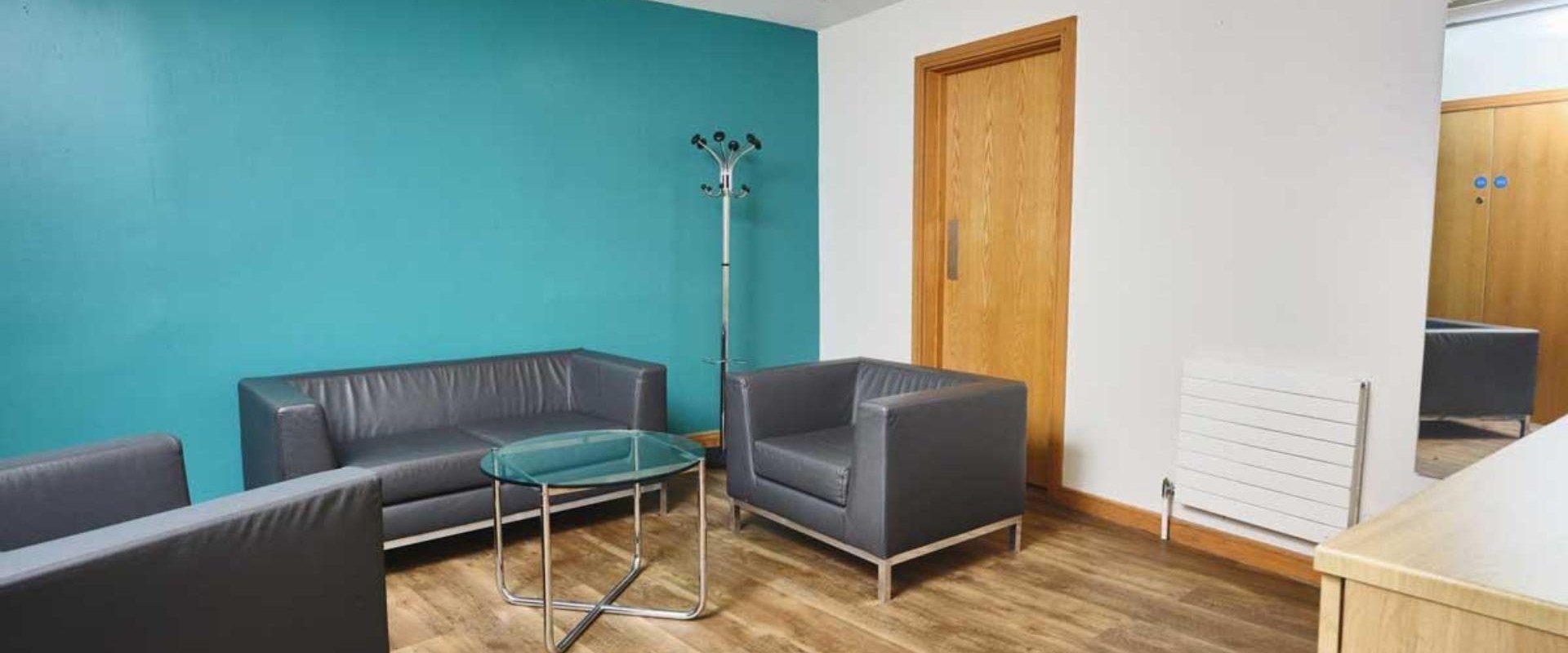 This image shows one corner of the Mezzanine Rooms at ICC Belfast with one blue wall and one white wall with a wooden door. Two grey sofas, a grey armchair and a coffee table are also in the room.