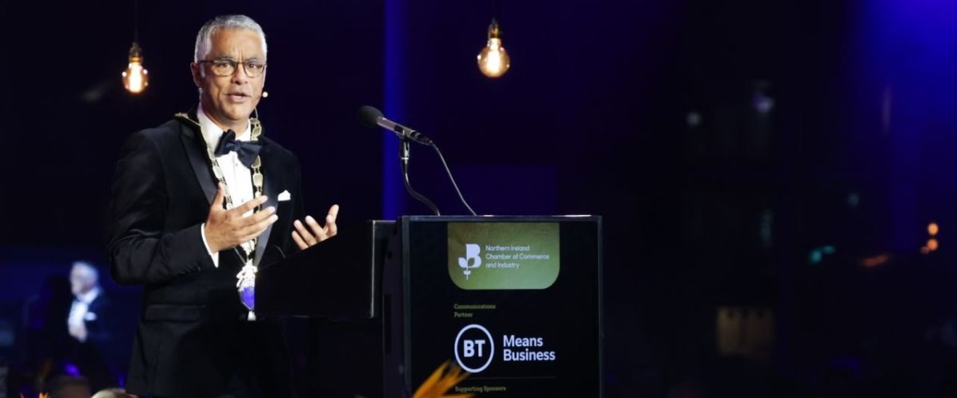 Man in Presidential Chains addresses audience in a gala dinner setting