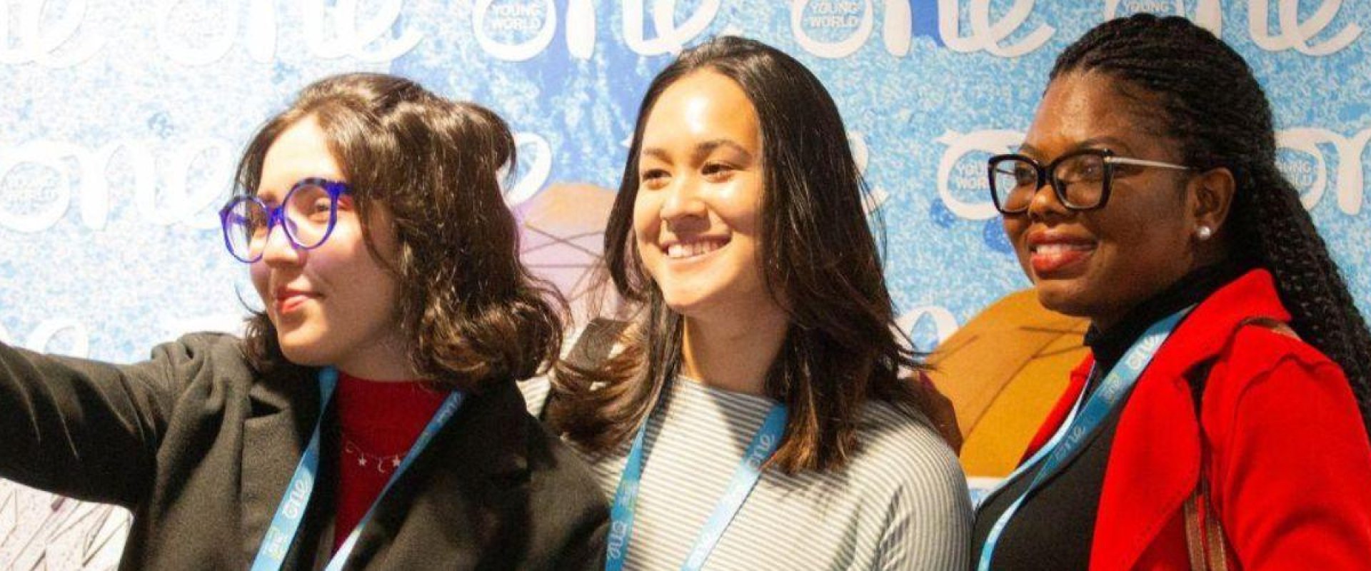 Three One Young World Delegates gathered in front of a branded backdrop in Hall 1, taking a group selfie with an iPhone.