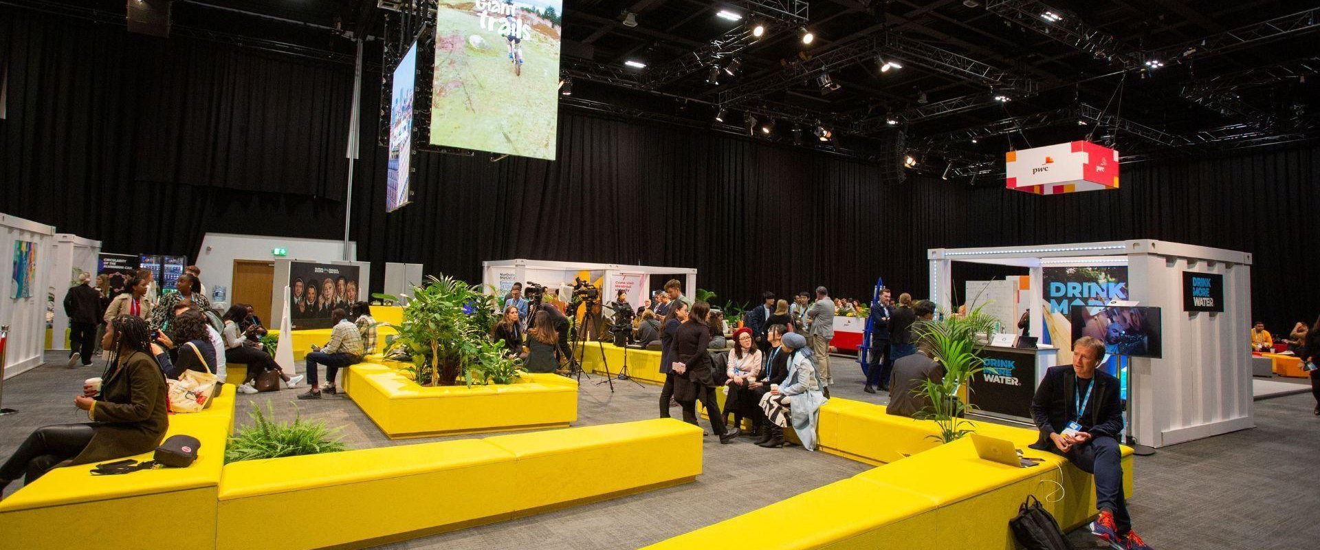 Men and women sitting in the exhibition space with long yellow seating, greenery and digital screens.