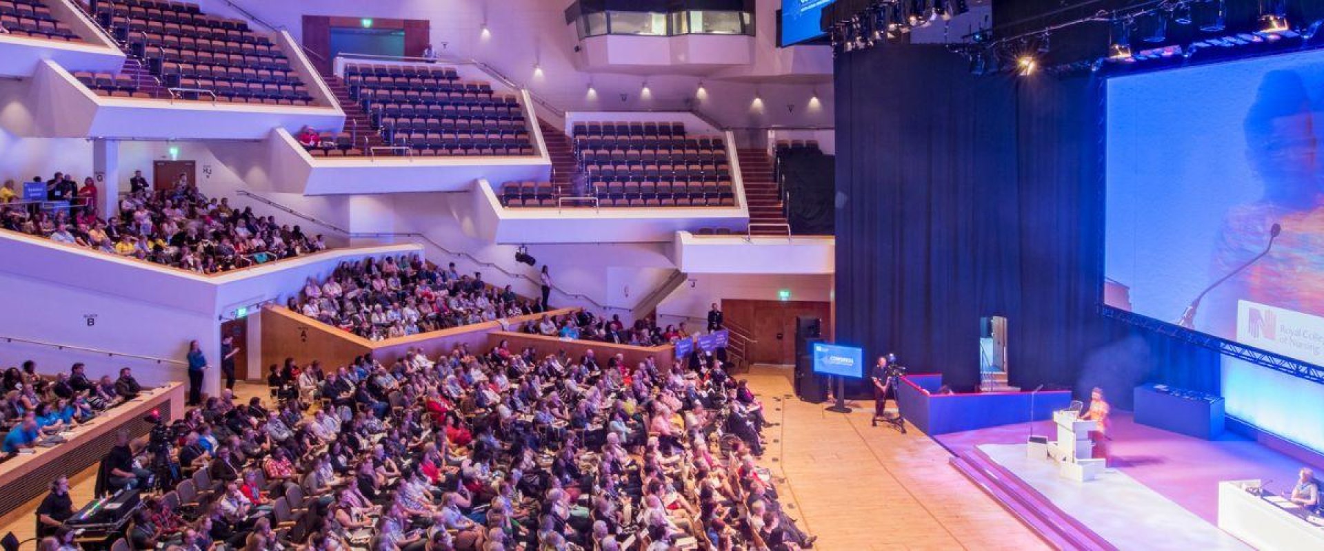 Main auditorium filled with people sitting in seats facing a stage which has a speaker and panel of four people sitting behind a desk in front of a big screen