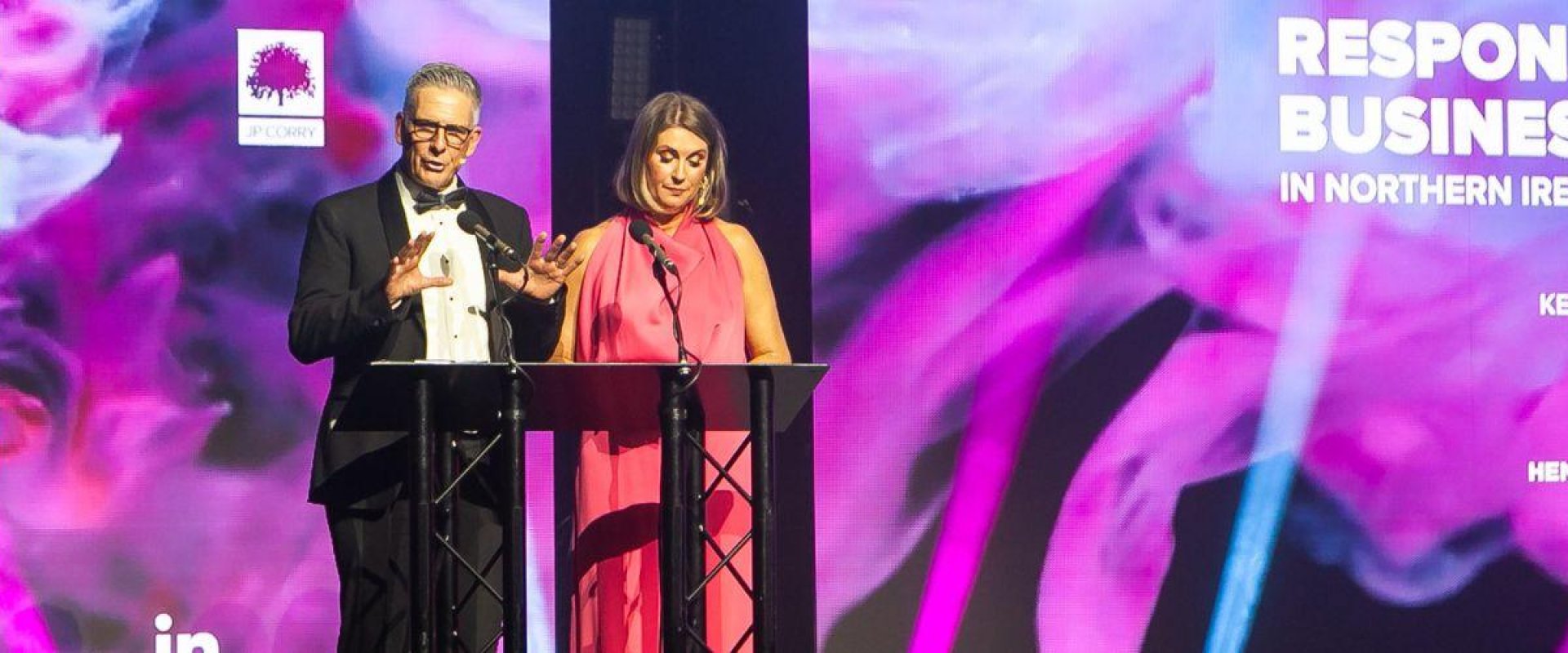A woman and man stage on a stage in front of a podium with a large purple digital screen behind them with the word 'Responsible Business Awards' on display