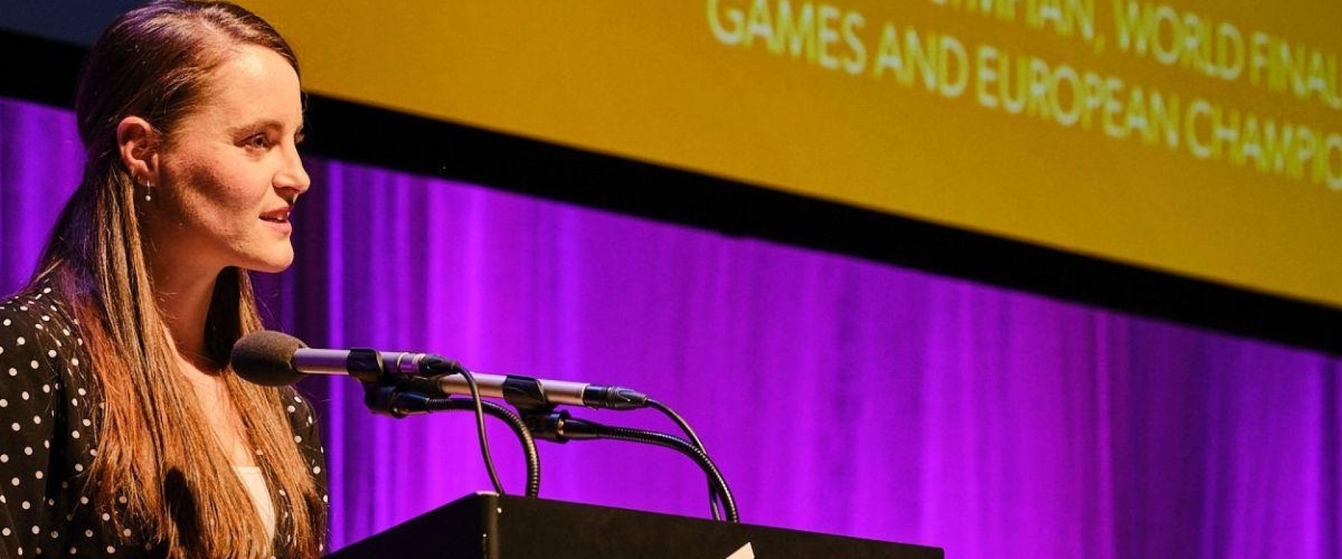 Ciara Mageean speaking at the SistersIN Event in 2023. She is stood on stage at a podium with a microphone, behind her is a curtain light up by purple lights and a yellow screen.