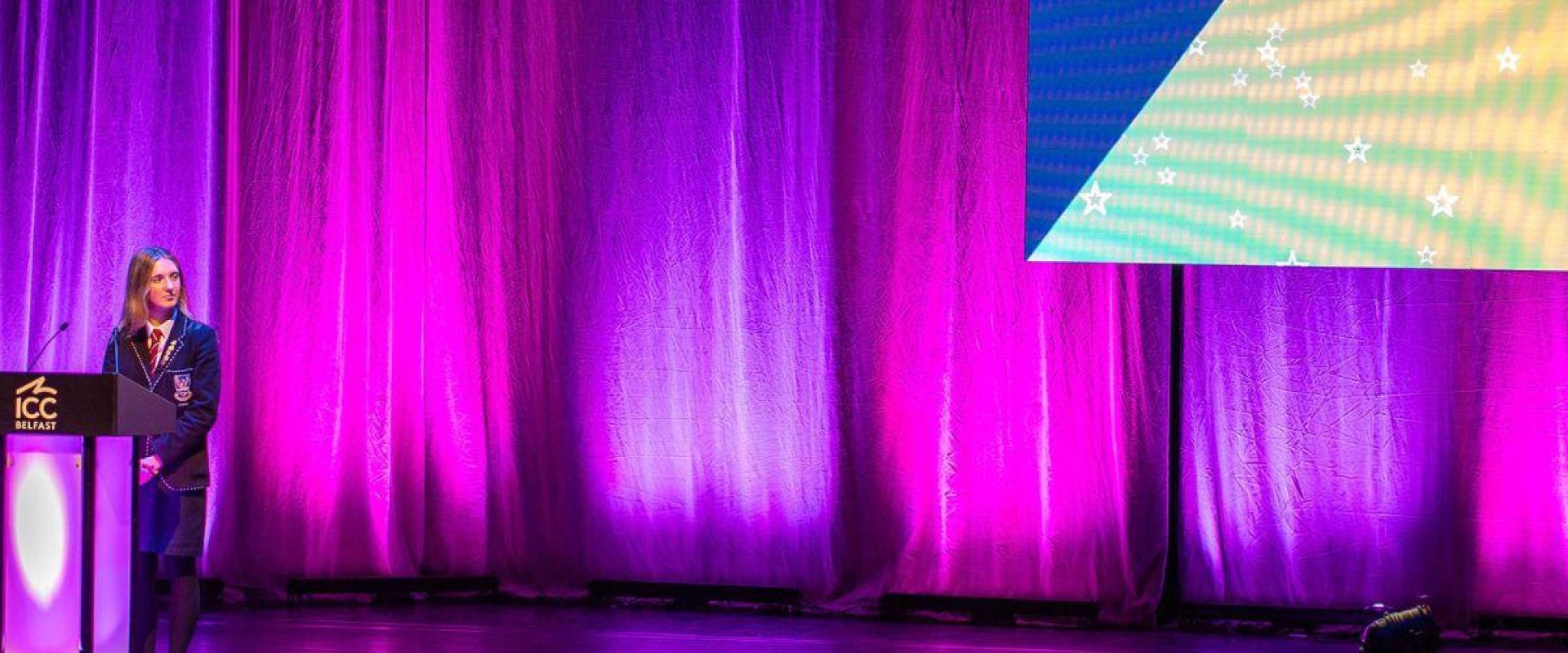 Two SistersIN pupils from the 2024 cohort on stage in the main auditorium, standing behind podiums as they gave their speeches. The curtains behind them shine purple and pink.