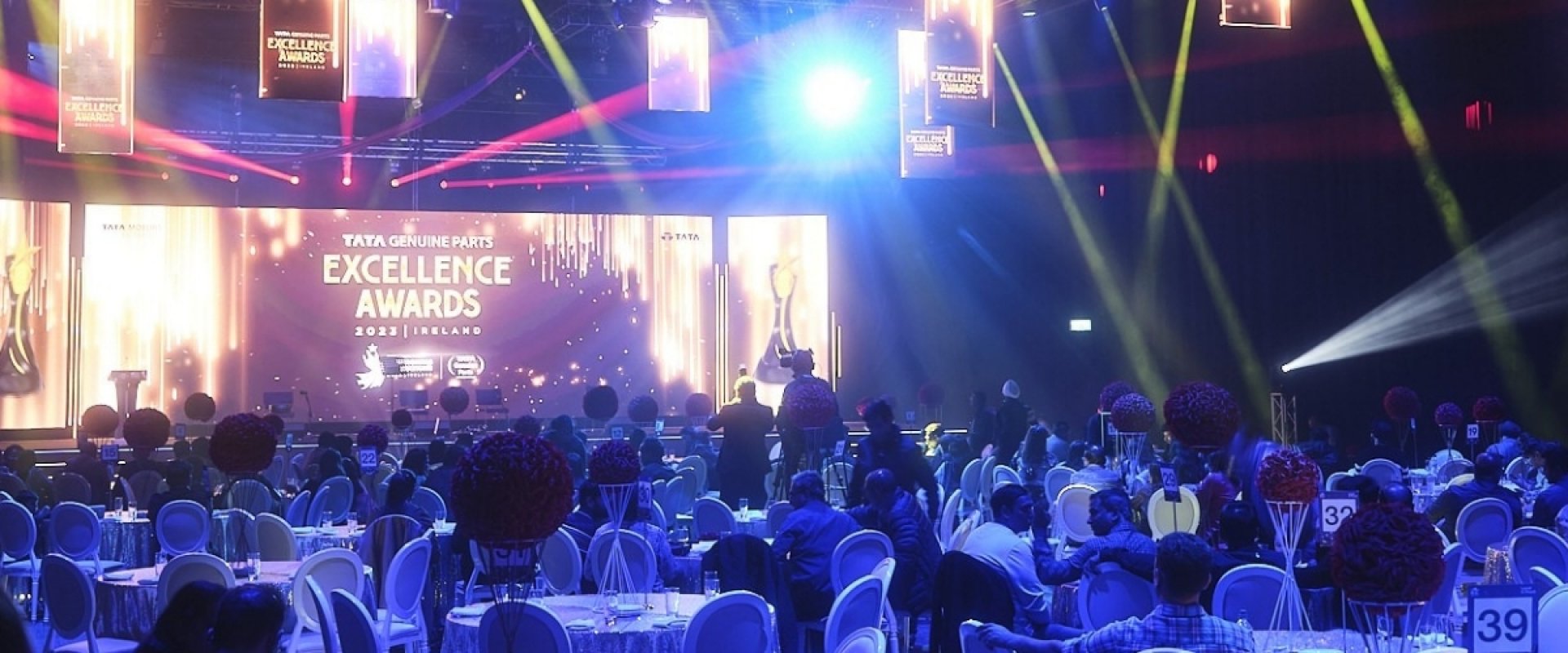 Hall 1 set up for the TATA Genuine Parts (TGP) Annual distributor conference award ceremony with round tables set up in the back facing a stage with coloured lights and a large digital screen that reads TATA Genuine Parts Excellence awards.