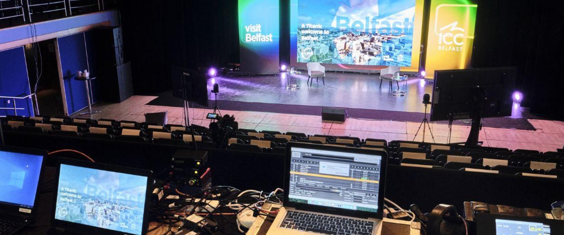 Image shows a production studio with multiple digital devices on display looking onto a large digital screen with the words 'Visit Belfast' visible. ICC Belfast hybrid event venue with live streaming and in-person audience.