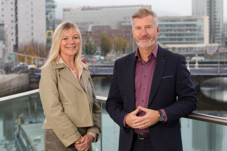 Two people standing outdoors on a balcony overlooking a river and cityscape. The woman on the left is wearing a light beige jacket and smiling, while the man on the right is wearing a dark suit with a maroon shirt and also smiling. Buildings and a bridge are visible in the background.