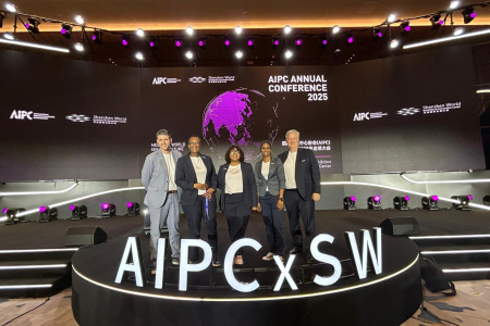 Four adults stand on a stage which displays illuminated letters AIPC X SW. It it two men and two women, all in trousers and jackets.