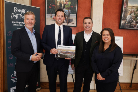 Three men in suits and a lady in black stand in front of a wall painted orange that has hanging paintings. One man in the centre is holding a brochure which has the words Draft Belfast City Airport Master Plan 2040