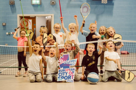 Image of children on a tennis court indoors cheering for press shot. Some are holding tennis rackets as props meanwhile one in the middle is holding a prop to mark the achievement