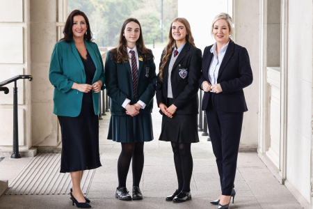 Two women in formal attire standing either side of two female students dressed in uniform stand proudly smiling for a press shot