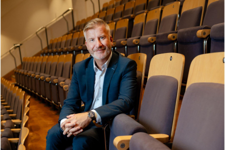 Man in his fifties in a navy suit and blue shirt sitting in an auditorium with his hands together looking at the camera. Behind him are rows of empty theatre seats.