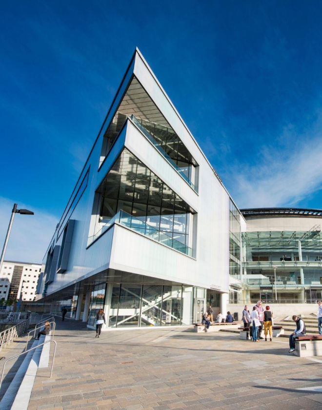 White pointed building which shows three levels, two with a balcony and to the side is a curved building with windows. Multiple adults standing and sitting outside the building in the sunshine.