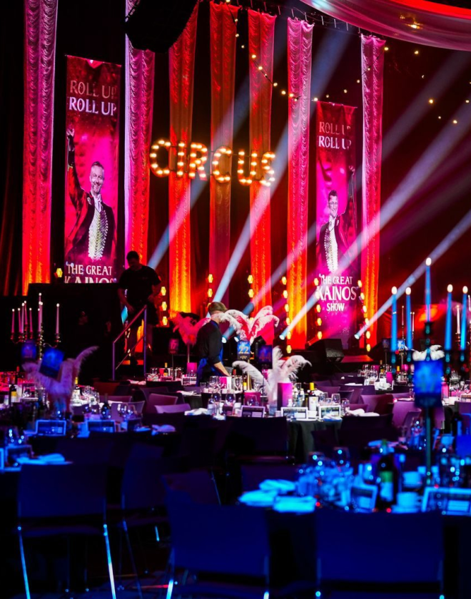 Hall filled with circular tables and chairs facing a stage that has a hanging banner displaying the words 'Circus' in yellow against a red background
