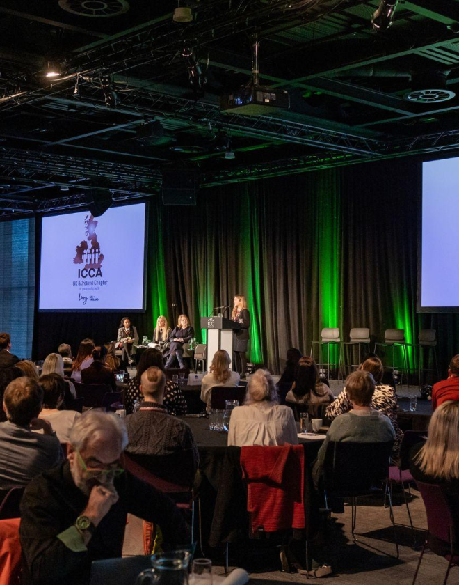 Students present on stage at the ICCA UK & Ireland Chapter to a packed hall full of people sitting at round tables on chairs