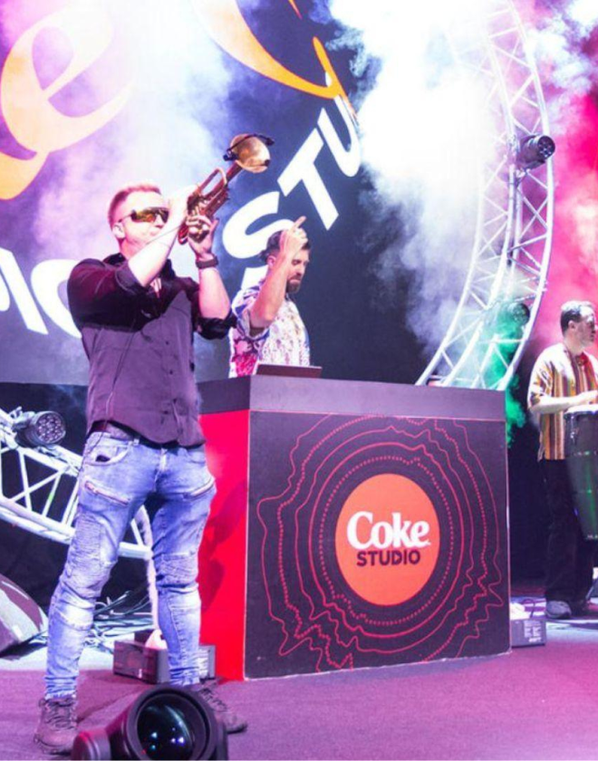 Man playing a trumpet on a stage in Hall 1 with a DJ playing in front of decks branded Coca-Cola. Digital signage also shows the words 'Coke Studio'