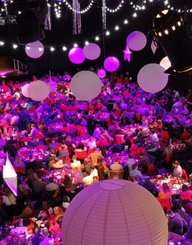 Image shows more than thirty tables full of people at a Christmas party in ICC Belfast. There is a stage in the background and lantern decorations and strings of lights hanging from the ceiling.