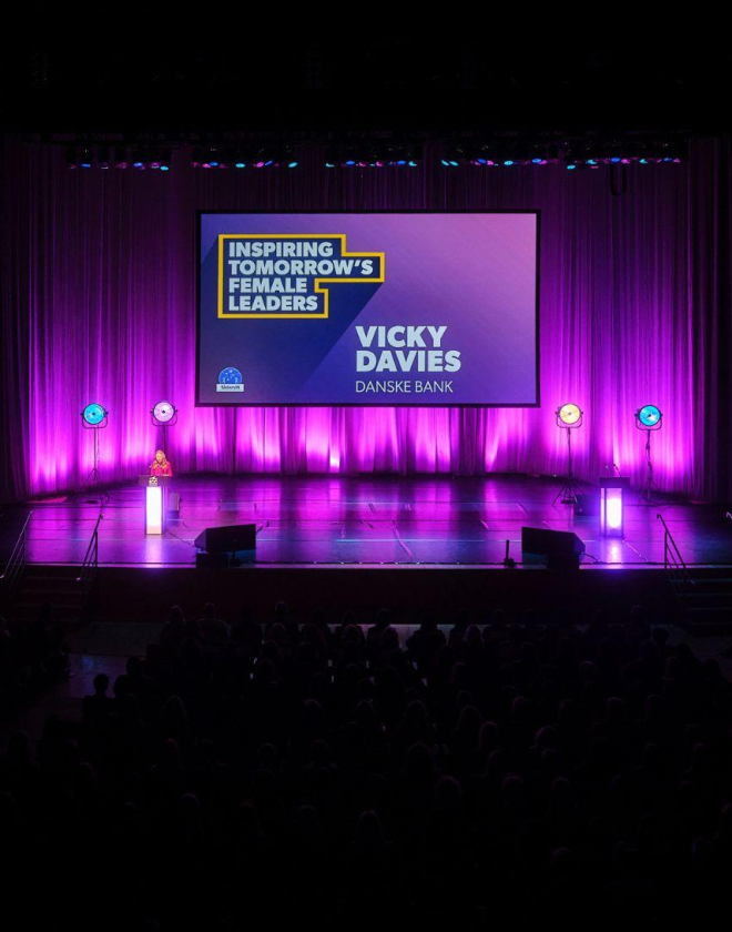 Main Auditorium set up for the SistersIN celebration event. Vicky Davies is stood at a podium delivering a presentation to the attendees and is stood in front of a large digital screen that displays her name and reads, ' Inspiring tomorrow's female leaders'