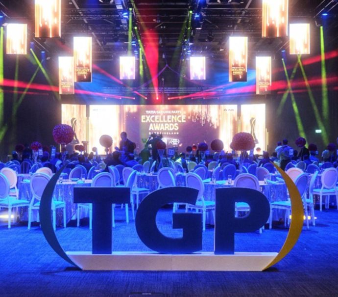 Hall 1 set up for the TATA Genuine Parts (TGP) Annual distributor conference award ceremony, large branding statue that reads (TGP) with round tables set up in the back facing a stage with coloured lights and a large digital screen that reads TATA Genuine Parts Excellence awards.