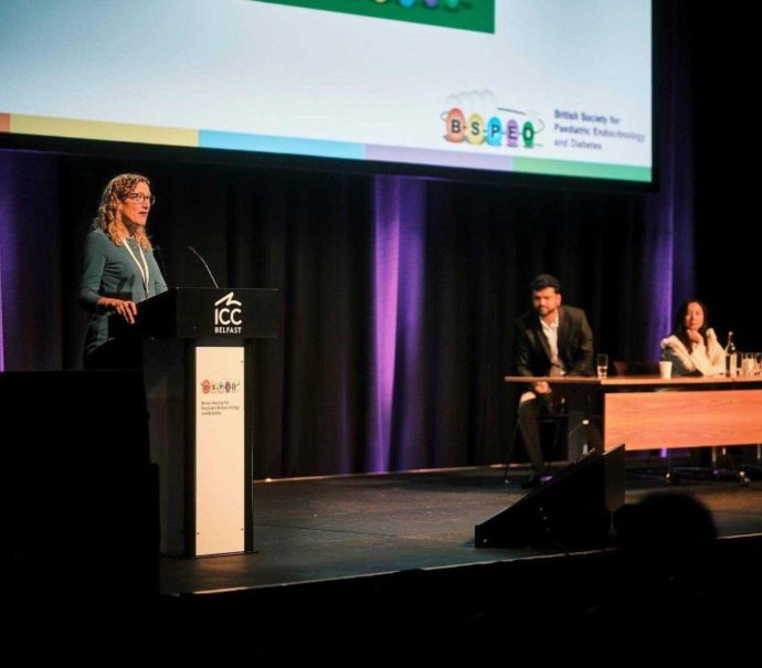 Tabitha Randell, president of BSPED, addresses people from podium on stage with two members of her team in the background sitting at a table