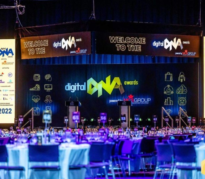 Image displays a large room set up for an awards dinner. At the front of the room is various lit up signs for the Digital DNA Awards.