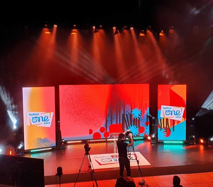 The ICC Belfast Studio stage set up for the hybrid launch of One Young World 2023. Large screens are on stage lit by red lights showing a graphic with the One Young World Belfast logo.
